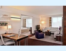 Helles, modernes Hotelzimmer mit separatem Wohn- und Schlafbereich. Im Vordergrund ein Schreibtisch mit Stuhl, daneben ein Wohnzimmer mit Sofas, Sessel, Couchtischen und einem Fernseher; im Hintergrund ein Bett mit großen Fenstern und Vorhängen.