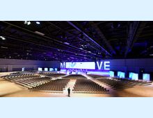 Großer Konferenzsaal mit einer Bühne im Hintergrund, beleuchtet in Blau und Lila, und einer großen Bestuhlung in Reihen. Die Halle hat eine hohe Decke mit sichtbaren Trägern und Beleuchtung, sowie eine Videoleinwand hinter der Bühne.