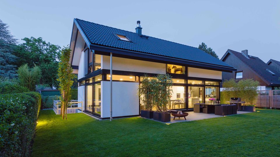 Modernes Einfamilienhaus mit Satteldach und großer Glasfront, beleuchtet in der Dämmerung. Im Vordergrund liegen ein gepflegter Rasen und eine Terrasse mit Outdoor-Möbeln, umgeben von Hecken und Nachbarhäusern.