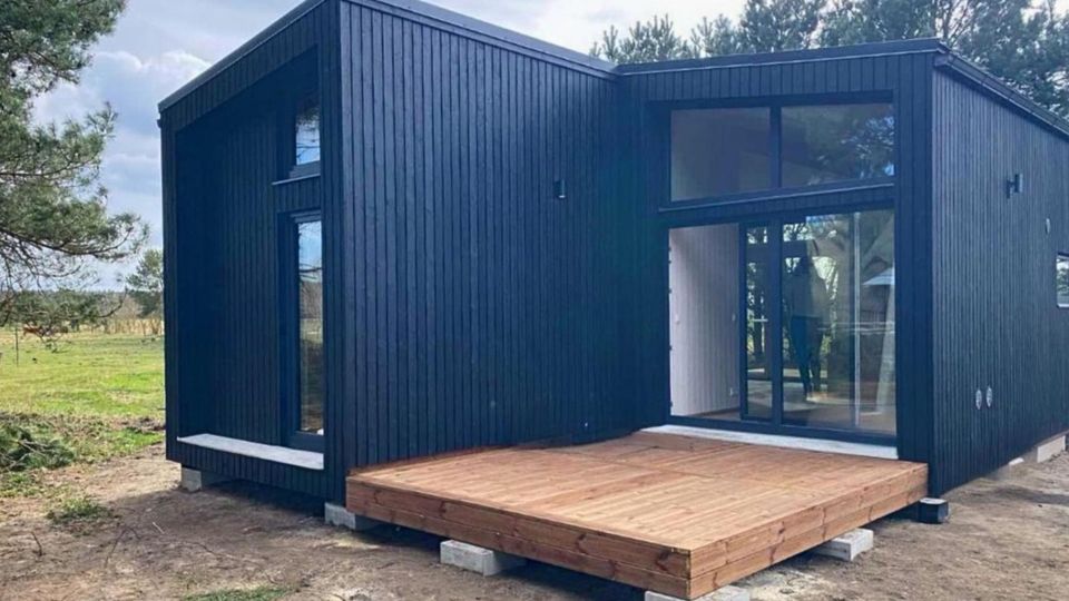 Modernes, kubisches Tiny House mit schwarzer Holzfassade und asymmetrischem Pultdach, davor eine Holzterrasse auf Betonfundamenten. Große bodentiefe Glasfronten und schmale Fenster, gelegen auf einer offenen Wiese mit Bäumen am Rand.