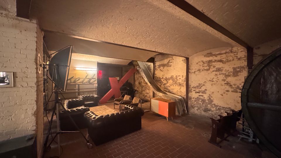 Industrieähnlicher Raum mit freiliegenden Ziegelwänden, Gewölbedecke und rotbraunem Fliesenboden. Im Vordergrund steht eine Sitzgruppe mit dunklen Ledersofas um einen Holztisch, im Hintergrund befinden sich ein großes rotes "X", eine silberne Rutsche sowie verschiedene Möbel und Lampen.