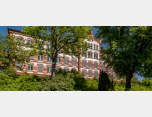Backsteingebäude im Stil der Gründerzeit mit weißen Fensterrahmen und einem Schriftzug „Burgel Schule“ auf der Fassade, umgeben von hohen Bäumen und dichtem Grün. Der klare Himmel deutet auf sonniges Wetter hin.