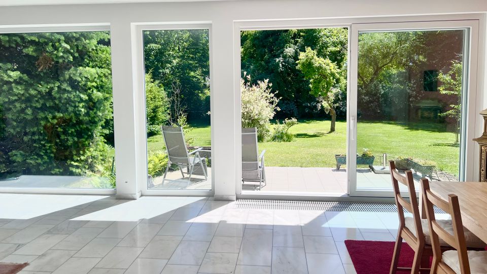 Heller Wohn- und Essbereich mit großflächigen bodentiefen Fenstern und einer Schiebetür, die auf eine geflieste Terrasse führen. Draußen ist ein großer, grüner Garten mit Rasen, Bäumen und Sträuchern zu sehen.