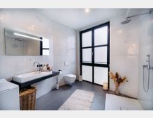 Modernes Badezimmer mit großen, undurchsichtigen Fenstern mit schwarzem Rahmen, einer Wand aus hellen Fliesen, einer offenen Regendusche und einem Waschbecken mit Spiegel. Der Raum ist minimalistisch eingerichtet mit einem Teppich, dekorativen Elementen wie einem Korb und getrockneten Pflanzen in einer Vase.