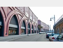 Straßenansicht eines Gebäudes mit roten, großen Rundbogenfassaden, in denen Schaufenster und Türen eingebaut sind. Links und rechts der Straße befinden sich geparkte Autos, und moderne Straßenlaternen säumen den Gehweg.