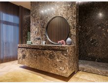 Luxuriös gestaltetes Badezimmer mit Wänden und Waschtisch aus poliertem braunem Marmor, einem runden Spiegel, eingebautem Wasserhahn und hölzernen Elementen. Am linken Bildrand befinden sich vertikale Holzlamellen vor einem Fenster, und auf dem Boden liegt ein rund geformter gelber Teppich.