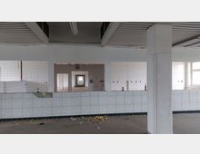 Ehemaliger gewerblicher Raum mit gefliesten Wänden und Boden, weißen Wandfliesen und Deckenplatten. Der Raum wirkt verlassen, mit sichtbaren Schäden und liegengebliebenem Schutt.