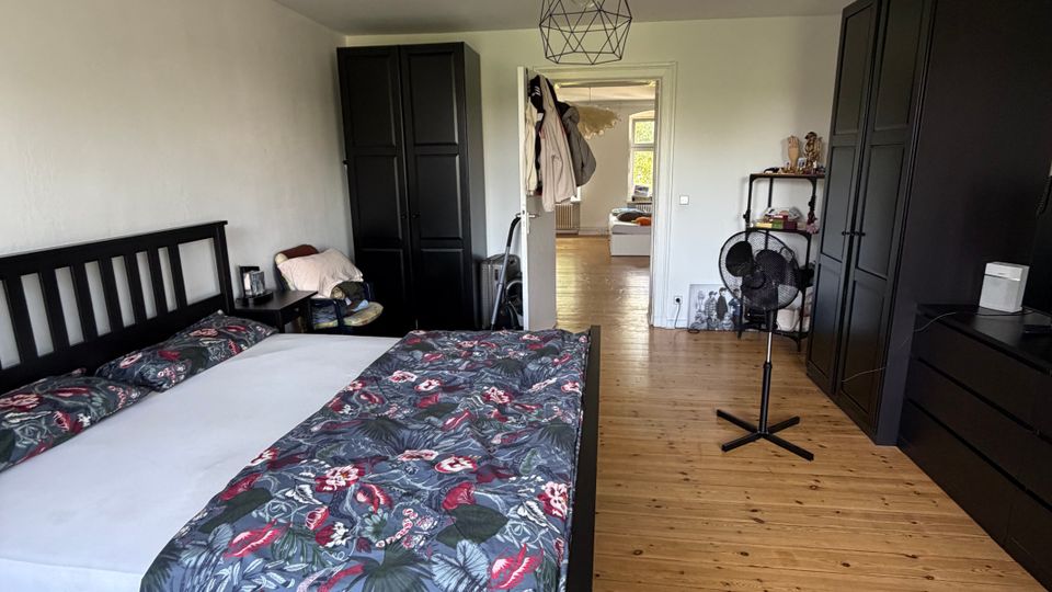 Schlafzimmer mit Holzdielenboden, großem Doppelbett mit dunklem Holzrahmen und gemusterter Bettwäsche. An den Wänden stehen mehrere schwarze Kleiderschränke; in der Mitte hängt eine geometrische Deckenlampe, und eine offene Tür führt in einen hellen Flur.