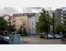 Straßenszene in einer städtischen Wohngegend mit mehrstöckigen Wohnhäusern; auf der linken Seite ein gelbes Gebäude mit Balkonen, daneben ein graues modernes Gebäude. Im Vordergrund ist eine Straßenkreuzung mit einem Vorfahrt-gewähren-Schild sowie geparkte Autos, Fahrräder und Bäume sichtbar.