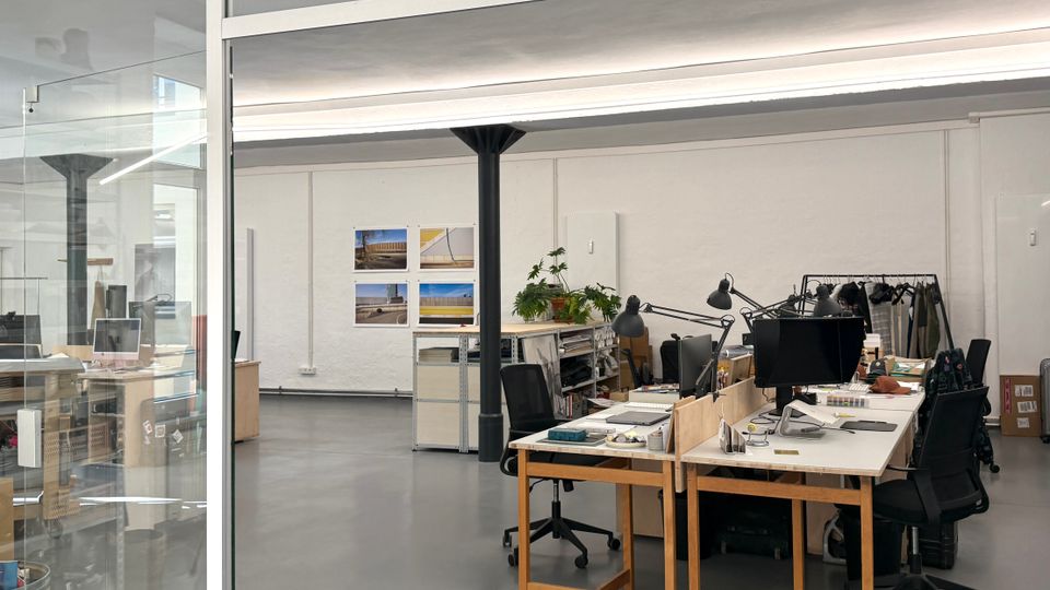 Heller Großraumbüro- oder Atelierraum mit grauem Betonboden, weißen Wänden und langen Deckenleuchten, in der Mitte von einer schwarzen Stützsäule getragen. Mehrere Schreibtische mit Monitoren und Arbeitslampen stehen neben Regalen, Pflanzen und einer Kleiderstange; links ist eine Glaswand und oben ein grünes Notausgangsschild.