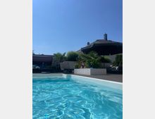 Ein moderner Außenbereich mit einem rechteckigen Pool, von dem ein Teil des glänzenden, blauen Wassers zu sehen ist. Im Hintergrund befinden sich Liegestühle, Sonnenschirme, Pflanzen in Pflanzkästen und ein Gebäudedach unter klarem, blauem Himmel.