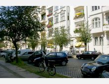 Straßenszene mit einer Reihe von mehrstöckigen Altbauhäusern mit verzierten Fassaden und Balkonen, auf denen Pflanzen und Sonnenschirme zu sehen sind. Im Vordergrund parkende Autos auf Kopfsteinpflaster und ein Fahrrad an einem Baum.