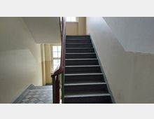 Treppenhaus mit grauen Stufen, einem Holzgeländer und hellbeigen Wänden. Im Hintergrund befindet sich ein Fenster, das natürliches Licht einfallen lässt.