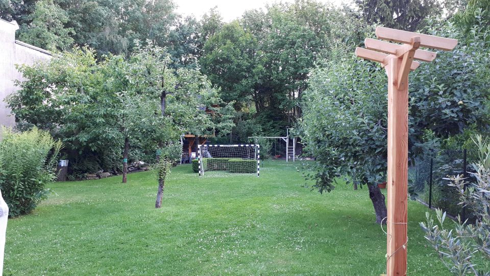 Großer, gepflegter Garten mit Rasenfläche, Obstbäumen und dichtem Baumbestand am Rand; in der Mitte steht ein kleines Fußballtor. Rechts im Vordergrund befindet sich ein Holzpfosten mit Querbalken, daneben Sträucher und ein Zaun.
