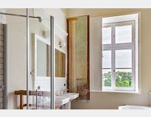 Helles Badezimmer mit großer Fensterfront, durch die Grünflächen im Außenbereich sichtbar sind. Das Badezimmer verfügt über zwei Waschbecken mit Wandspiegeln, eine Duschkabine mit Glaswand sowie weiße Wandfliesen und eine dekorative Wandmalerei mit Säulenmotiv.