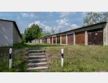 Reihe von Garagen mit unterschiedlich farbigen Holztoren, umgeben von einer grünen Wiese und Bäumen im Hintergrund. Im Vordergrund befinden sich betonierte Treppenstufen und zwei schwarz-weiß markierte Pfosten.