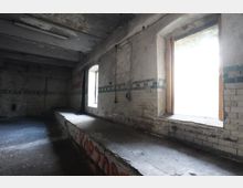 Verlassener Industrie- oder Lagerraum mit stark abgeplatztem Putz, bröckelnder Decke und einer halbhohen gefliesten Wand mit grünem Fliesenband. Zwei hohe Fensteröffnungen lassen grelles Tageslicht herein; entlang der Wand verläuft eine lange, erhöhte Betonplattform mit Graffiti.