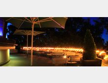 Terrasse eines Restaurants bei Nacht, beleuchtet von warmem gelben Licht entlang eines Geländers, mit Holzbänken und Tischen sowie großen Sonnenschirmen; im Hintergrund dunkle Bäume und ein tiefblauer Himmel.