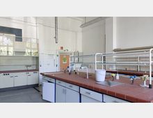 Ein Chemielabor mit einem langen Arbeitstisch aus roten Fliesen, ausgestattet mit Wasserhähnen, Spülbecken und Gasbrennern. Im Hintergrund sind eine Abzugshaube, Schränke und eine Tür zu sehen; die Wände sind weiß und mit Leitungen und Regalen versehen.