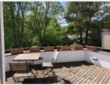 Holzterrasse mit einem kleinen Tisch und zwei Stühlen, umgeben von einer weißen Brüstung, auf der Blumentöpfe stehen. Im Hintergrund sind grüne Bäume und ein heller Himmel sichtbar.