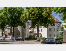 Straßenszene mit mehrstöckigen Wohnhäusern im klassischen Stil, hell verputzt und mit roten Ziegeldächern. Vor den Gebäuden stehen Bäume, geparkte Autos und Laternen; ein Straßenschild weist auf die "Brentanostraße" hin.