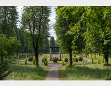 Ein parkartiger Garten mit hohen Bäumen, einer zentralen Kiesweg-Achse und einer überdachten Steinpergola in der Mitte. Im Hintergrund sind symmetrisch angelegte Wege, Büsche und Rankgerüste zu sehen.