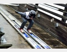 Ein Skispringer in grauem Anzug und schwarzem Helm befindet sich in der Anlaufspur einer hölzernen Sprungschanze, bereit zum Absprung. Die schmale Spur ist mit Schienen und Werbeaufklebern versehen.