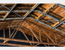 Dachkonstruktion mit sichtbaren Holzbalken und Metallträgern, die eine Holzdecke stützen. Die Struktur ist offen und weist diagonale Verstrebungen sowie bauliche Details auf.