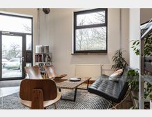 Ein gemütlicher Warteraum mit modernen Möbeln, darunter zwei braune Holzstühle, eine schwarze Ledercouch und ein Holztisch auf einem grauen Teppich. Im Raum stehen Pflanzen, ein Regal mit Büchern und eine große Fenster- und Glastürfront, durch die Tageslicht einfällt.