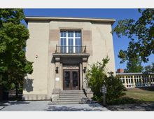 Ein Kulturhaus mit heller Putzfassade und symmetrischer Architektur. Der Eingang besteht aus einer Doppeltür mit Glas- und Metallelementen, erreichbar über eine kurze Treppe, flankiert von Laternen; daneben befinden sich Bäume und ein moderner Anbau im Hintergrund.