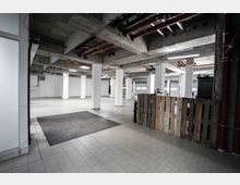 Industriehalle mit rohbelassenem Beton an Decke und Pfeilern, hellen Fliesenboden und sichtbaren Rohrleitungen an der Decke; ein Bereich ist mit einer Absperrung aus Holzpaletten versehen.