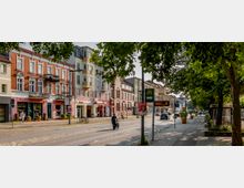 Straßenansicht einer deutschen Stadt mit mehrstöckigen Wohn- und Geschäftshäusern, teils mit Backsteinfassaden und Balkonen. Entlang der breiten Straße mit Gehwegen stehen Bäume, unter denen sich Sitzmöglichkeiten und Laternen befinden; eine Bushaltestelle und Straßenschilder sind ebenfalls sichtbar.