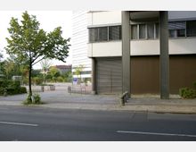 Moderne Gebäudefassade mit grauen Stützen und einem geschlossenen Rolltor, dahinter eine gepflasterte Fläche mit Bäumen und einem Parkbereich. Im Vordergrund ein Bürgersteig, eine Straße und ein einzelner Baum.