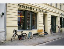 Fassade eines Geschäftes mit der Aufschrift „Whisky & Cigars“ auf einem hellgelben Gebäude mit grünen Fensterrahmen. Vor dem Laden stehen ein Fahrrad, eine Holzbank und eine geschriebene Tafel auf dem Gehweg.