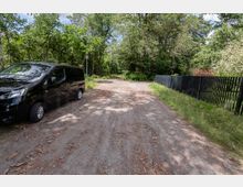 Unbefestigter Waldweg mit breiter, sandiger Fahrspur, umgeben von dichtem Grün und hohen Bäumen. Rechts verläuft ein schwarzer Lattenzaun entlang des Weges, im Hintergrund steht eine einzelne Straßenlaterne.