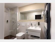 Modernes Badezimmer mit einer gläsernen Duschkabine, einem wandmontierten WC und einem weißen Waschtisch mit Spiegel. Die Wände sind mit hellen Fliesen verkleidet, und an der rechten Seite befindet sich ein Handtuchheizkörper.