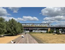 Eine städtische Parklandschaft mit einer Holzplattform und Sandfläche im Vordergrund, umgeben von Bäumen. Im Hintergrund fährt eine gelbe U-Bahn über eine erhöhte Stahlbrücke mit Graffiti an den darunterliegenden Wänden, unter einem blauen Himmel mit vereinzelten Wolken.