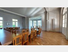 Großer, lichtdurchfluteter Raum mit langen Holztischen und passenden Stühlen auf einem Fischgrätenparkettboden. Zwei Wände sind von großen Fenstern durchbrochen, die viel Tageslicht hereinlassen, und ein angrenzender Erkerbereich enthält zusätzliche Sitzgelegenheiten.