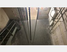 Blick in einen kleinen, industriellen Raum mit unverputzten Backsteinwänden, einer Metalltreppe links, Ketten, die von der Decke hängen, und freiliegenden Rohren an der Wand. Der Boden ist aus dunklem Material, und die Beleuchtung ist gedämpft.