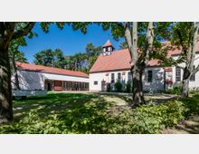 Weiß verputzte Kirche mit rotem Ziegeldach und kleinem Glockenturm, daneben ein modernes Gemeindezentrum mit großen Fenstern und Flachdach, umgeben von Bäumen und einer Grünfläche. Im Hintergrund sind Wald und blauer Himmel sichtbar.