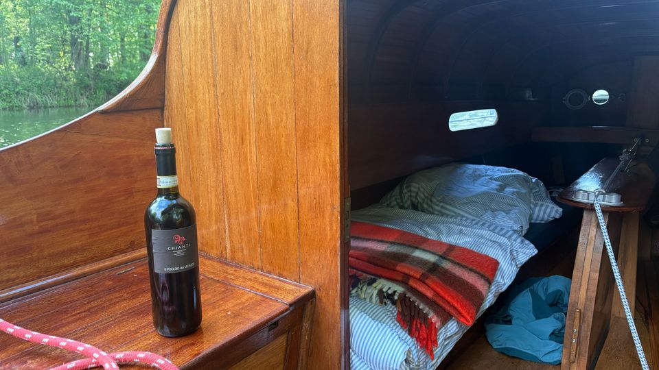 Blick in die Schlafkajüte eines kleinen Holzbootes mit dunkel getäfeltem, gebogenem Innenraum und schmalen Fenstern. Im Vordergrund steht auf einer Holzbank eine Weinflasche neben einem Seil; in der Kajüte liegen Bettzeug und Decken, seitlich ist ein kleiner Klapptisch zu sehen.