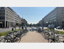 Straßenszene in einer urbanen Umgebung mit modernen Gebäuden links und rechts; im Vordergrund Fahrradständer mit geparkten Fahrrädern, mittig ein Eingang zur U-Bahn-Station mit einem „U“-Schild, dahinter eine breite Straße mit Fußgängern, Autos und Fahrrädern. Im Hintergrund ist der Berliner Fernsehturm sichtbar.