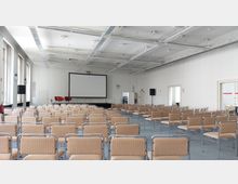 Konferenzraum mit mehreren Reihen beige gepolsterter Stühle, einem Beamer-Leinwand an der Vorderseite und zwei roten Sesseln auf einer kleinen Bühne. Der Raum hat helle Wände, eine Rasterdecke mit integrierter Beleuchtung und große Fenster auf der linken Seite.