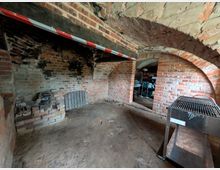 Alter Backstein-Kellerraum mit gewölbter Decke, sichtbar beschädigten Wänden und schwarzer Verfärbung durch Ruß. Im Hintergrund befindet sich ein Metallgrill und Regale mit Gegenständen.