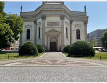 Außenansicht einer historischen Kirche mit klassizistischer Fassade, zentralem Portal, hohen Fenstern und mehreren Statuen auf dem Dach. Der Vorplatz besteht aus einem gepflasterten Weg mit symmetrischem Muster, umgeben von gepflegten Büschen und Grünflächen.