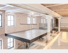 Industrieller Innenraum mit Backsteinwänden, großen, bogenförmigen Fenstern und einer im Bau befindlichen Glaswand für einen Konferenzraum. Der Raum verfügt über sichtbare Stahlträger, weiße Säulen und ein Zwischengeschoss mit Geländer.