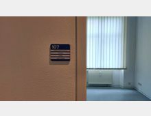 Wand mit einem Türschild, auf dem „107 Gesundheitsamt“ und weitere Informationen stehen, daneben ein Raum mit hellem Boden, einer weißen Heizung und bodenlangen Lamellenjalousien vor dem Fenster.