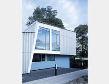 Modernes Gebäude mit klaren geometrischen Formen, verkleidet mit Wellblechpaneelen und großen Glasfenstern. Der obere Bereich ist weiß und grau, während der untere in einem hellblauen Farbton gehalten ist; umgeben von Kiesflächen, mit Gehwegplatten im Vordergrund und Bäumen im Hintergrund.