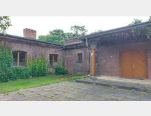 Ziegelgebäude mit flachem Dach und begrünten Mauern, teilweise bewachsen mit Efeu. Im Vordergrund befindet sich ein gepflasterter Hof, daneben ein kleiner Grünbereich; rechts ist ein großes Holztor und links mehrere Fenster mit Holzrahmen sichtbar.