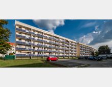 Ein mehrstöckiges Wohngebäude mit Balkonen in Blau-Weiß; davor befindet sich eine Wiese, ein kleiner Parkplatz mit Autos und ein rotes Auto im Vordergrund. Der Himmel ist teils bewölkt, und es gibt Bäume an den Seiten des Gebäudes.